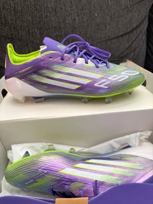 Adidas F50 elite  lila/gröna fotbollsskor - Säljer ett par Adidas F50 elite fotbollsskor i en riktigt snygg lila och grön färgkombination med vita detaljer. Skorna har klassisk snörning, låg profil och är tillverkade i ett lätt syntetmaterial för maxad speed på planen. Perfekta för dig som vill sticka ut och spela snabbt.