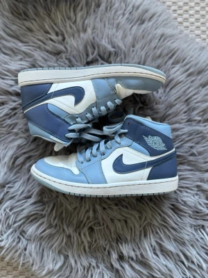 Nike Air Jordan 1 Mid blå/vit - Snygga Nike Air Jordan 1 Mid sneakers i blått och vitt läder med klassisk swoosh-logga på sidorna och Air Jordan Wings-märke vid ankeln. Skorna har perforerad tå, blå snörning och vit sula. Perfekta för dig som gillar streetwear och ikonisk design.