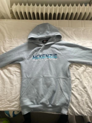Ljusblå hoodie från McKenzie - Ljusblå hoodie från McKenzie med broderad logga i blått och vitt på bröstet. Klassisk känguruficka framtill och huva. Mjuk bomullsblandning som är skön att ha på sig. Perfekt för en avslappnad stil.