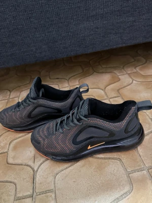 Nike Air 720 svarta sneakers - Nike Air 720 sneakers i svart mesh med orange detaljer och synlig Air-sula. Skorna har en sportig design med rundad tå, snörning och reflekterande detaljer. Perfekta för dig som gillar streetwear och vill ha en bekväm och snygg sneaker.