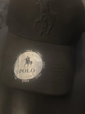 Svart keps från Polo Ralph Lauren - Snygg svart keps från Polo Ralph Lauren med broderad logga framtill och justerbar rem baktill. Kepsen är i klassisk baseballmodell och har detaljer med Polo-tryck på insidan. Perfekt accessoar för en sportig och stilren look.