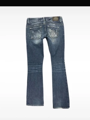 Blå bootcut jeans från Guess - Snygga blå bootcut jeans från Guess med slitningar och broderad detalj på bakfickan. Jeansen har glitter/stenar på backfickan, låg midja och lätt tvättad look. 