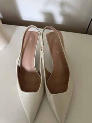 Vita spetsiga pumps från mei miu - Snygga vita pumps från mei miu med spetsig tå och öppen häl. Skorna har en elegant slingback-design och är tillverkade i ett glansigt material som ger en lyxig känsla. Perfekta för att lyfta din outfit med en stilren touch.
