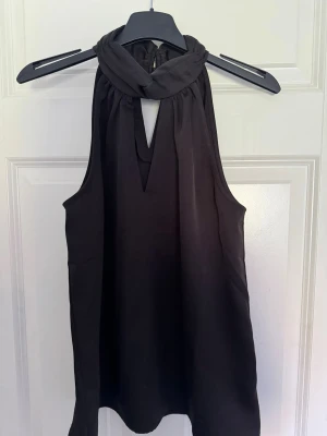 Svart ärmlös halterneck topp - Snygg svart ärmlös topp med halterneck och twistad krage. Toppen har en liten v-ringad öppning framtill och ett elegant, flowy fall. Perfekt för utgång!