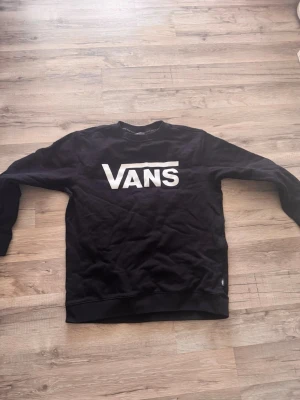 Svart sweatshirt från Vans - Svart sweatshirt från Vans med stor vit logga framtill. Klassisk rund halsringning och långa ärmar. Perfekt för en avslappnad streetstyle-look. Mjuk bomullskänsla och ribbade muddar vid ärmslut och nederkant.