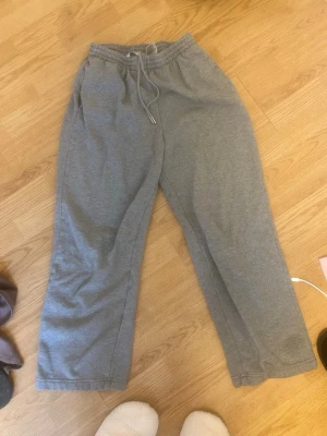 Gråa mjukisbyxor med dragsko - Ett par klassiska grå mjukisbyxor med resår i midjan och justerbar dragsko. Byxorna har raka ben och är tillverkade i mjukt bomullsmaterial. Perfekta för en avslappnad stil.