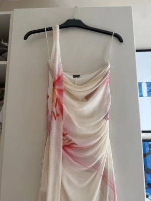 Vit och rosa långklänning  - Elegant långklänning i vitt med rosa blommiga detaljer. Med en tillhörande lång scarf. Klänningen har draperad front. Tunt, flowy material som ger en romantisk känsla. Perfekt för sommarens speciella tillfällen.