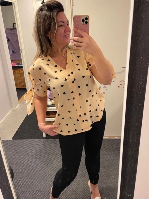 Beige blus med svarta hjärtan - Söt beige blus med svarta hjärtan och V-ringning. Blusen har korta ärmar med knytband på sidorna som ger en extra detalj. Luftigt och lätt material, perfekt för en trendig och avslappnad look.helt ny