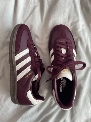 Adidas Samba vinröda sneakers - Adidas Samba sneakers i vinrött med klassiska vita ränder och detaljer. Ovandel i läder och mocka, gummisula och guldfärgad SAMBA-text på sidan. Snygg retrostil med låg profil och ikonisk design som passar till många outfits.