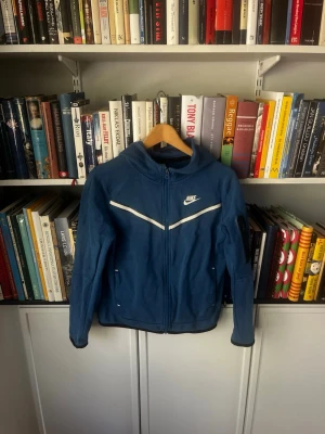 Blå tech fleece från Nike med dragkedja - Snygg blå vindjacka från Nike med vit detalj över bröstet och hel dragkedja framtill. Jackan har hög krage, långa ärmar och två fickor med dragkedja. Perfekt för en sportig och avslappnad stil.