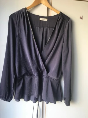 Mörkblå omlottblus från 365 - Snygg mörkblå omlottblus från 365 med v-ringning och knytband i midjan. Blusen har långa ärmar och ett lätt, flowigt material som ger en avslappnad och trendig look. Perfekt att matcha med jeans eller kjol för en cool stil.
