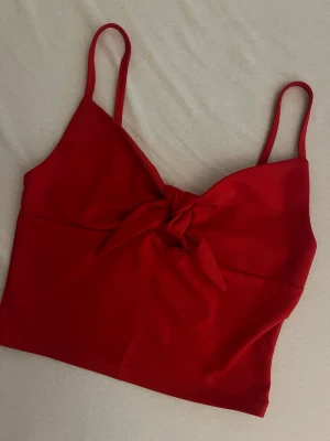Röd crop top med knut framtill - Snygg röd crop top med tunna axelband och en dekorativ knut framtill. Toppen har en figurnära passform och är gjord i ett mjukt, stretchigt tyg som känns bekvämt mot huden. Säljer för 40kr. 