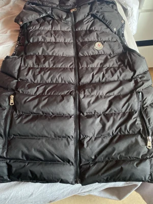 Svart dunväst från Moncler - Snygg svart dunväst från Moncler med dragkedja framtill och två fickor med dragkedja. Västen har Moncler-logga på bröstet och diskreta detaljer längs axelbanden. Perfekt för lager-på-lager och håller dig varm under kyliga dagar. Priset kan diskuteras.