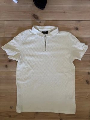 Vit kortärmad topp med dragkedja - Vit topp från Primark med korta ärmar och strukturmönster. Toppen har en liten krage och en snygg dragkedja framtill som detalj. Perfekt för en stilren och fräsch look.