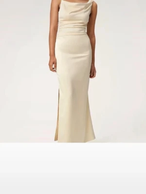 Gul/Champagnefärgad långklänning i satin - Elegant långklänning i champagnefärgad satin med draperad urringning och slits på sidan. Klänningen har figurnära passform och är ärmlös, perfekt för speciella tillfällen. Snygg och stilren design som ger en lyxig känsla.