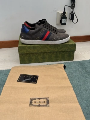 Gucci Ace sneakers med GG-mönster - Gucci Ace sneakers i mörkgrått med klassiskt GG-mönster och guldfärgade detaljer. Skorna har blå och röda ränder på sidan samt kontrasterande hälflikar i rött och blått. Vit sula och svarta skosnören. Lyxig och ikonisk design i canvas och läder.