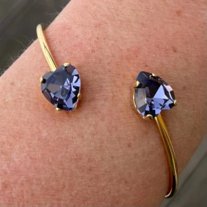 Armband, Bangle, guldpläterad hjärt kristaller.  - Armband, Bangle, i guldpläterad mässing med kristaller. Hjärtformade kristaller 11x10mm. Färg Tanzanite. ställbar storlek. De senare bilderna andra armband som finns i lager. Jag skickar endast spårbart, samfraktar. Fast pris. Besök gärna mina övriga annonser.  Till mina smycken använder jag kristaller från Preciosa, Aurora och Swarowski.