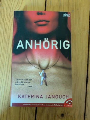 Anhörig av Katerina Janouch - En gripande roman av Katerina Janouch som utforskar livet som anhörig till en alkoholist. Boken har ett rött och beige omslag med ett konstnärligt motiv och tydlig titel. Pocketutgåva med extramaterial om författaren.