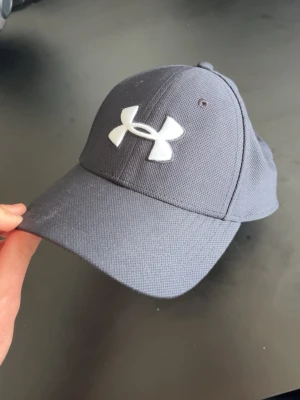 Mörkgrå keps från Under Armour - Snygg mörkgrå keps från Under Armour med böjd skärm och stort vitt logotryck framtill. Kepsen är tillverkad i ett slitstarkt tyg och har klassisk sexpanelsdesign med ventilerande hål. Perfekt för en sportig och avslappnad stil.