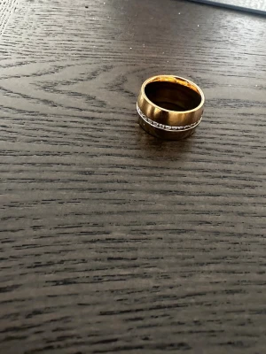Edblad ring  - Snygg bred ring i guldfärgad metall med en rad gnistrande stenar längs mitten. Ringen har en klassisk och stilren design som passar perfekt för dig som gillar att sticka ut. Perfekt accessoar för att lyfta din look.