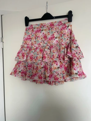 Blommig volangkjol i rosa toner - Supersöt kort kjol med blommigt mönster i rosa, vitt och grönt. Kjolen har dubbla volanger som ger en lekfull look och är perfekt för varma dagar. Tillverkad i ett lätt och luftigt material som känns skönt mot huden.