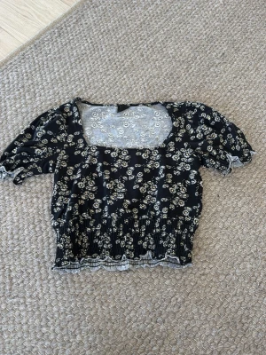 Svart blommig blus med puffärm - Säljer en svart blus med blommigt mönster och puffiga korta ärmar. Blusen har en fyrkantig halsringning och smockad nederkant med volangdetalj. Perfekt för dig som gillar romantiska och trendiga toppar.