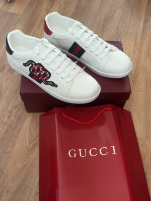 Gucci Ace vita sneakers med orm - Gucci Ace sneakers i vitt läder med klassiska gröna och röda ränder på sidan samt en broderad röd och svart orm. Skorna har vita snören, låg profil och detaljer i rött och svart på hälen. Perfekta för dig som vill sticka ut med lyxig streetstyle.Hör av er vid intresse