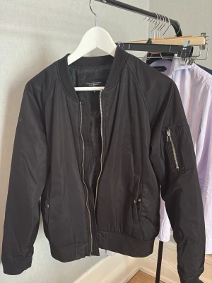 Svart bomberjacka från Zara Man - Stilren svart bomberjacka från lager 157  med klassisk passform, silverfärgad dragkedja och fickor med tryckknapp. Jackan har ribbade muddar vid ärmslut och midja samt en ärmficka med dragkedja. Perfekt för en trendig streetstyle-look.