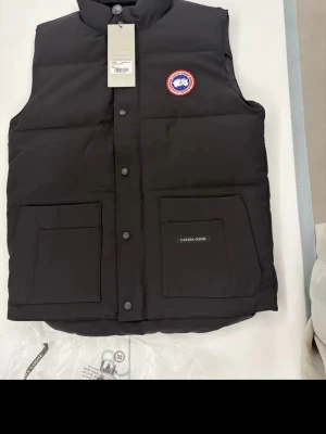 Svart dunväst från Canada Goose - Snygg svart dunväst från Canada Goose med klassisk logga på bröstet och två stora fickor framtill. Västen har hög krage, tryckknappar och är tillverkad i slitstarkt material med dunfyllning för extra värme. Perfekt för lager-på-lager och streetwear.