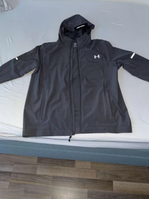 Svart vindjacka från Under Armour - Snygg svart vindjacka från Under Armour med huva och hel dragkedja. Jackan har två sidofickor med dragkedja och diskreta vita detaljer på ärmarna samt logga på bröstet. Perfekt för blåsiga dagar och sportiga outfits.