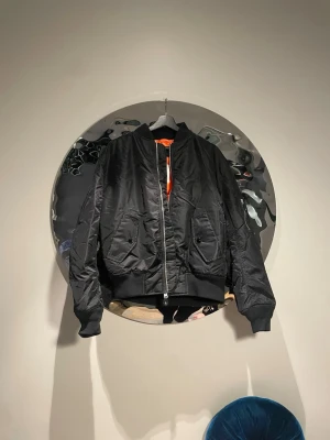 Ny Svart bomberjacka från hm  - Cool svart bomberjacka från hm  med klassisk orange foder. Jackan har två stora fickor med knappar, ribbade muddar och dragkedja framtill. Materialet är glansigt nylon och modellen är oversized med en rak passform.