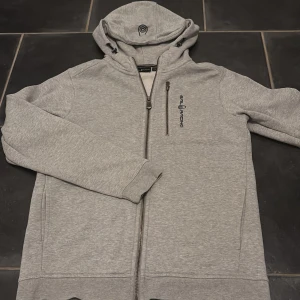 Ljusgrå hoodie från Sail Racing - Snygg ljusgrå hoodie från Sail Racing i storlek 170, med dragkedja framtill och en bröstficka med zip. Märkets logga är broderat på bröstet. Hoodien har huva med märket på. Hat bara använt 1 gång så i väldigt bra skick 