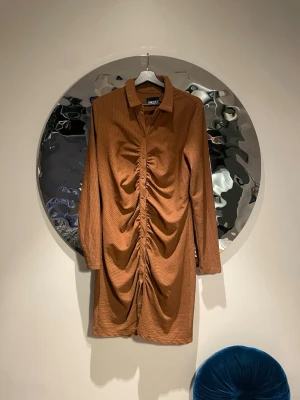 Brun ribbad klänning från PIECES - Snygg brun kortklänning från PIECES med ribbad struktur och lång ärm. Klänningen har en unik snörningsdetalj framtill som ger en cool rynkad effekt och knappar längs hela framsidan. Perfekt för dig som vill sticka ut med en trendig look.