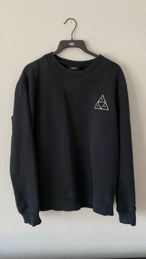 Svart sweatshirt HUF - Svart sweatshirt från HUF med klassisk crewneck och stort vitt triangellogotyp-tryck på ryggen samt mindre logga på bröstet. Tillverkad i mjukt bomullsmaterial och har ribbade muddar vid ärmslut och nederkant. Perfekt för en avslappnad streetwear-look.