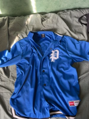 Blå baseball-inspirerad t-shirt - Snygg blå t-shirt med vita och svarta detaljer, inspirerad av baseballstil. Kortärmad med knappar framtill och ett stort vitt P på bröstet. Tillverkad i polyester och har sportig känsla med randiga muddar vid ärmslut och nederkant.