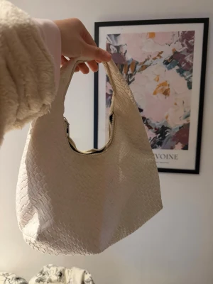 Vit handväska med ormmönster - Trendig vit handväska i konstläder med ormmönstrad struktur och rundad form. Väskan har ett kort handtag och dragkedja upptill. Perfekt accessoar för att lyfta din outfit med en cool detalj.