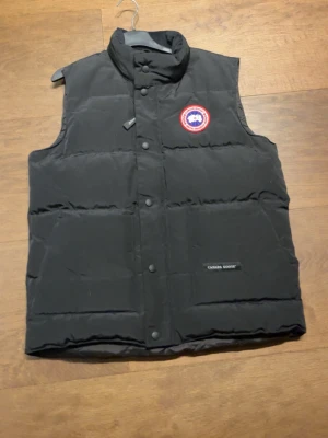 Svart dunväst från Canada Goose - Svart dunväst från Canada Goose med hög krage och knäppning framtill. Västen har ett klassiskt Canada Goose-märke på bröstet och en logotyp på ena fickan. Perfekt för kalla dagar och snygg att ha över en hoodie.