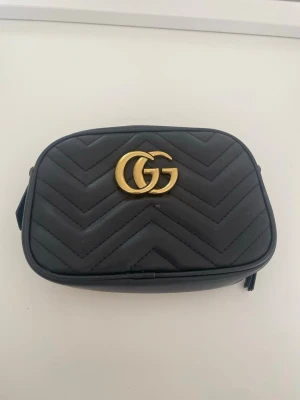 Svart  handväska i läder från  - Snygg svart handväska från  i quiltat läder med GG-logga i guld framtill. Väskan har en kompakt, rektangulär form och dragkedja i guld. Insidan är fodrad i beige tyg och har en liten innerficka. Perfekt för att bära det viktigaste.