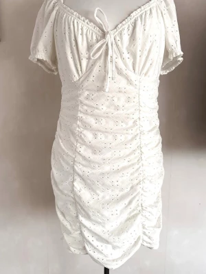 Vit kort klänning med puffärm - Söt vit kort klänning med puffärmar och broderade detaljer. Klänningen har omlottkänsla vid bysten med knytband och smockad resår vid ärmslut. Materialet är bomull med spetsmönster och lätt rynkad effekt längs sidorna.