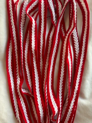 Rödvit dekorband accessoar - Säljer ett dekorband i textil med röd och vit färg. Bandet har ett fiskbensmönster och passar perfekt som accessoar till DIY-projekt, kläder eller väskor. Materialet är mjukt och flexibelt tyg, och bandet är smalt och långt. SKOSNÖREN!