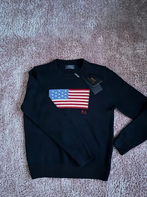 Mörkblå stickad tröja Polo Ralph Lauren - Snygg mörkblå stickad tröja från Polo Ralph Lauren med amerikansk flagga broderad på bröstet och RL-detalj i rött. Tröjan har rund halsringning och ribbade muddar vid ärmslut och nederkant. Perfekt för dig som gillar klassisk preppy stil.