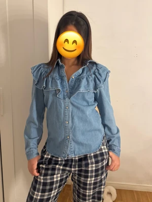 Blå jeansskjorta med volangdetaljer - Säljer en snygg blå jeansskjorta med volangdetaljer över axlar och bröst. Skjortan har långa ärmar, klassisk krage och knäppning framtill. Perfekt för dig som vill ha en trendig look