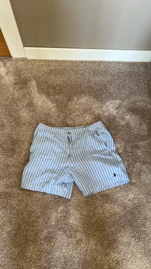 Blåvit randiga shorts från Polo Ralph Lauren - Säljer ett par blå shorts med vita vertikala ränder från Polo Ralph Lauren. Klassisk passform med knapp och dragkedja framtill samt fickor på sidorna. Ikoniska Polo-loggan broderad i mörkblått på benet. Perfekta för sommaren!