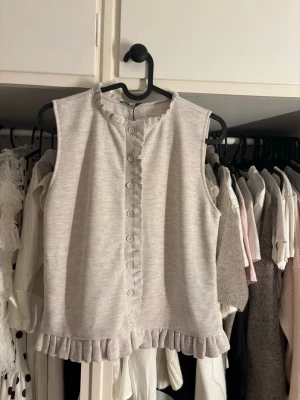 Beige ärmlös topp med volangkant - Supersöt beige ärmlös topp med volangkant längs nederkanten och knappar framtill. Toppen har en lätt ribbad struktur och en liten volang vid halsen. Aldrig använd och har prislapp kvar💕💕storleken är L men skulle definitivt säga att den sitter som en S