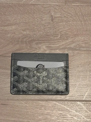 Grå korthållare - Snyggt och stilrent svart kortfodral med klassiskt mönster i vitt och grått. Fodralet har flera fack för kort och är gjort i exklusivt läder med präglad logotyp på framsidan. Perfekt för dig som vill ha en smidig och lyxig accessoar.