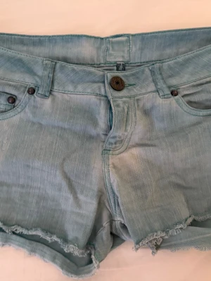 Ljusblå jeansshorts med fransig kant - Säljer ett par ljusblå jeansshorts från H&M med fransig kant nertill och klassisk femficksmodell. Shortsen har knapp och dragkedja framtill samt bälteshällor. Perfekta för varma dagar och enkel att matcha med olika toppar.