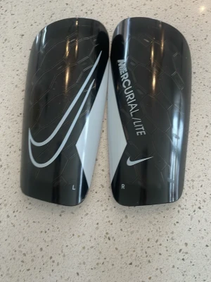 nike benskydd  - Säljer ett par Nike Mercurial Lite benskydd i svart med vita detaljer och stor Nike-logga. Lättviktsskydd med skön vadderad insida och ventilationshål för extra komfort. Perfekta för fotbollsträning eller match.