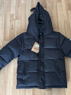 Svart pufferjacka från Burberry - Säljer en svart pufferjacka från Burberry med stor huva och dragkedja framtill. Jackan har två snedställda fickor med dragkedja och klassiskt rutigt foder i huvan. Perfekt för kalla vinterdagar och riktigt snygg streetstil.