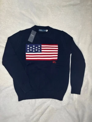 Mörkblå stickad tröja Polo Ralph Lauren - Mörkblå stickad tröja från Polo Ralph Lauren med stor amerikansk flagga på bröstet och RL-broderi i rött. Tröjan har rund halsringning och ribbade muddar vid ärmslut och nederkant. Perfekt för dig som gillar klassisk och stilren design. Ärmlängd:60 längd: 59 bröst: 48 axelbredd: 36