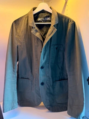 Olivgrön Cardigan Only & Sons - Snygg olivgrön kavajjacka från Only & Sons. Passar perfekt nu till våren! Storlek S. Det finns 2 svarta fläckar på jackan men syns knappt när man använder den. 
